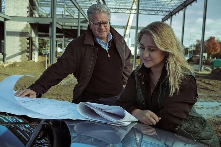 Two people reviewing blueprints on a car at a construction site with steel beams and dirt ground, under a cloudy sky, daytime.