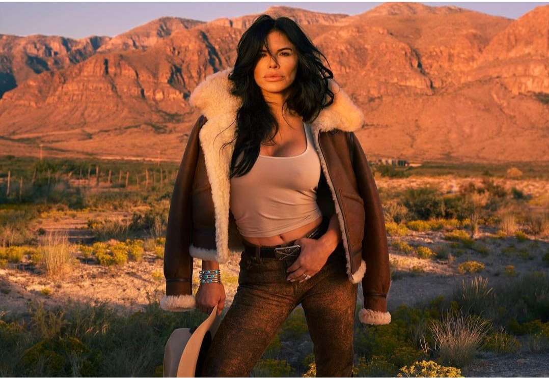 Brunette woman in shearling jacket posing outdoors in desert landscape with mountains at sunset wearing a tank top and patterned jeans.