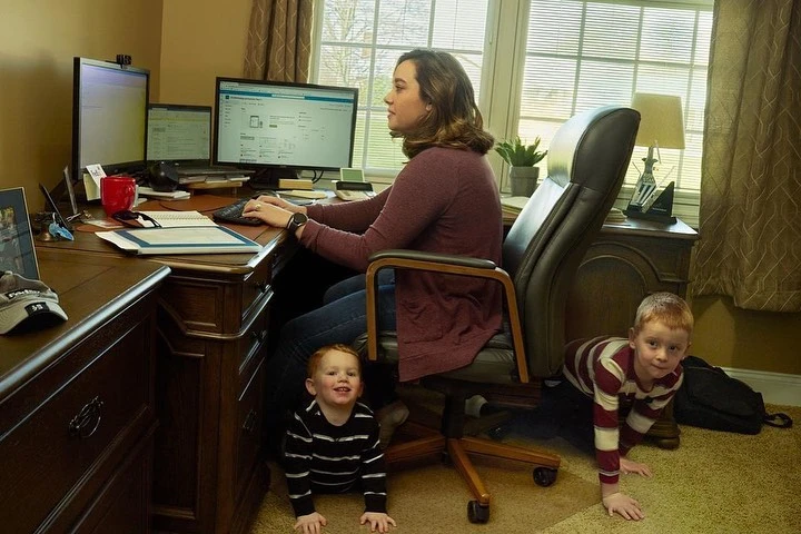 Woman works on desktop computer at home office while two young children play on the floor during the daytime in an indoor setting.