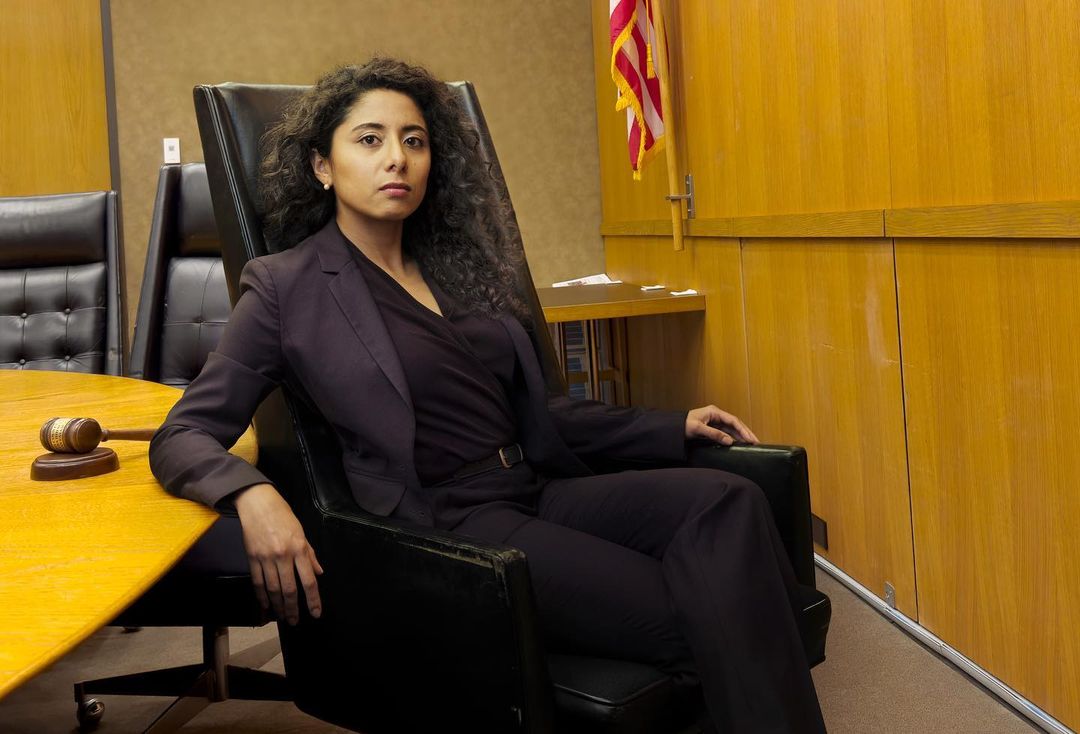 Woman in suit sits in courtroom chair with gavel on table. American flag and wood paneling in background. Focused portrait.