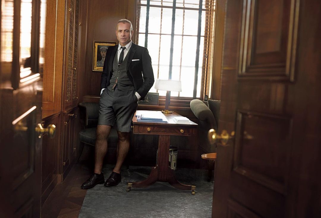Man in a suit with shorts posing indoors, leaning against a wooden desk with a lamp, chair, and framed artwork in the background.