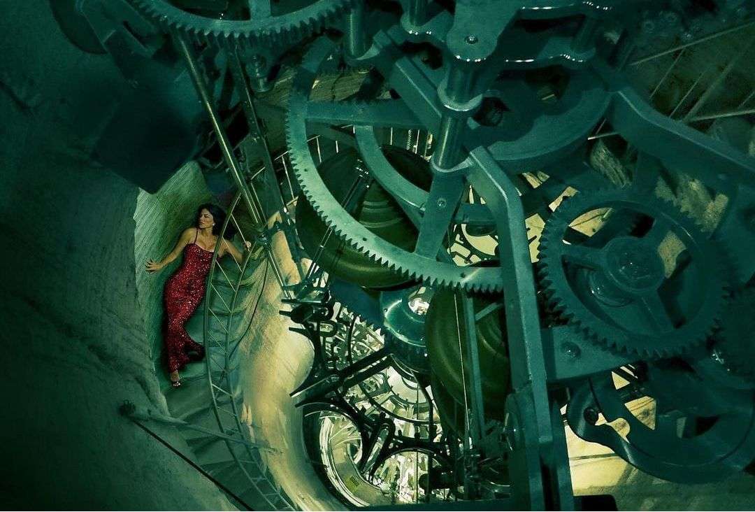 Woman in red dress poses on ladder amid giant clock gears inside a tower.  Complex machinery fills the background, teal and grey tones.