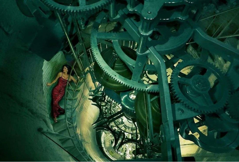 Woman in red dress poses on ladder amid giant clock gears inside a tower.  Complex machinery fills the background, teal and grey tones.
