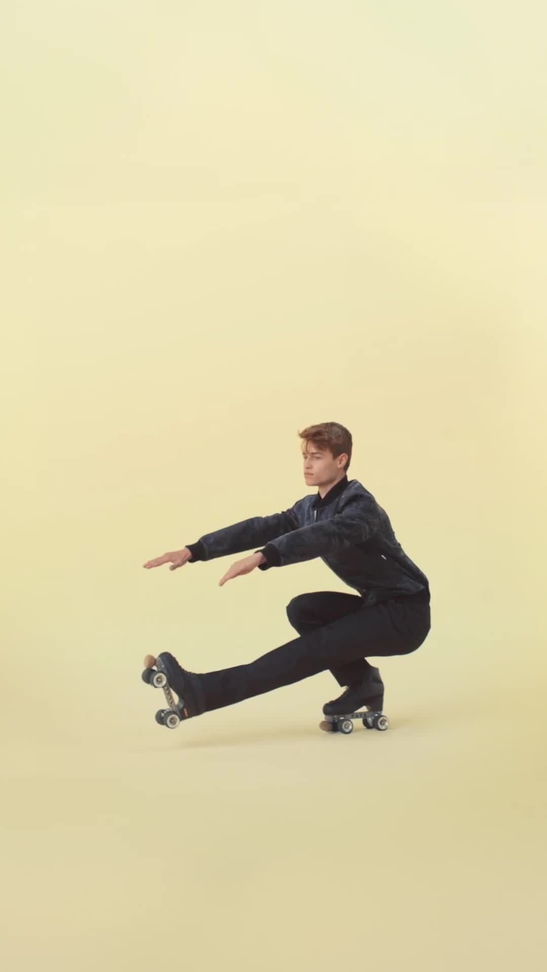 A person in a dark tracksuit and roller skates balances in a low squat against a pale yellow backdrop, extending their arms for balance.