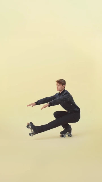 A person in a dark tracksuit and roller skates balances in a low squat against a pale yellow backdrop, extending their arms for balance.
