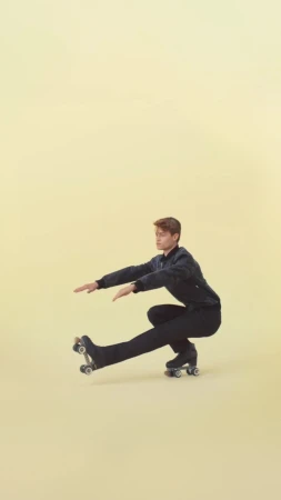 A person in a dark tracksuit and roller skates balances in a low squat against a pale yellow backdrop, extending their arms for balance.