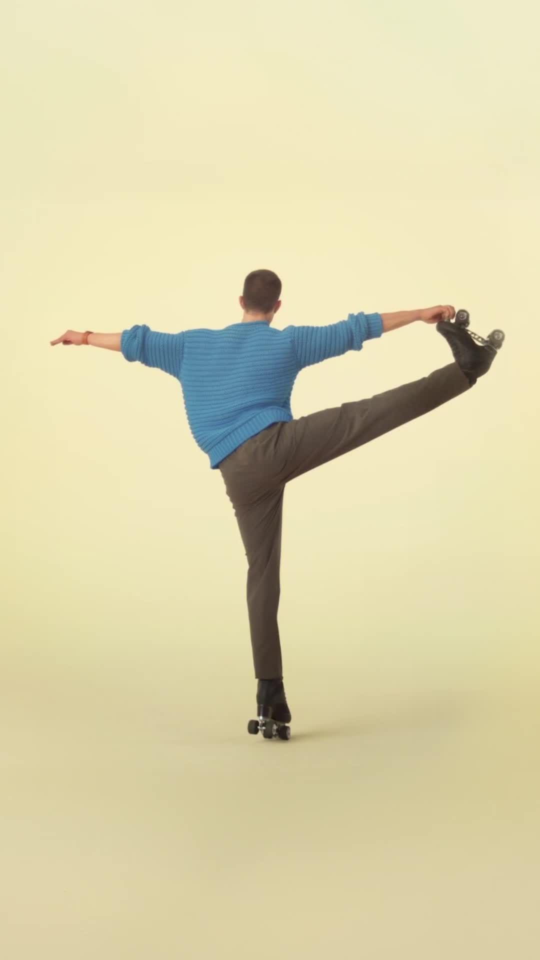 Adult male in gray pants and blue sweater performs a standing skating pose on roller skates in a studio setting with a yellow backdrop.