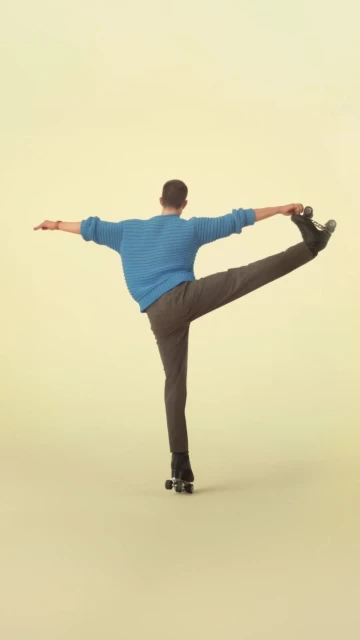 Adult male in gray pants and blue sweater performs a standing skating pose on roller skates in a studio setting with a yellow backdrop.