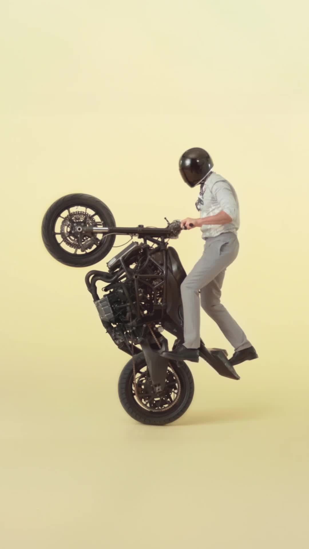 Person in helmet performing a motorcycle wheelie on a black motorcycle against a yellow background in a studio shot.