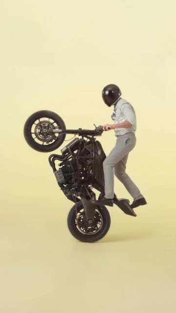 Person in helmet performing a motorcycle wheelie on a black motorcycle against a yellow background in a studio shot.