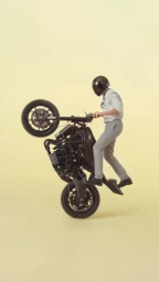 Man wearing a black helmet, light shirt, and grey trousers performing a motorcycle wheelie on a seamless neutral background.
