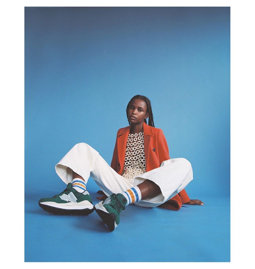 A fashion shot features a woman seated against a blue backdrop, wearing a red blazer, white pants, and green sneakers with colorful socks.