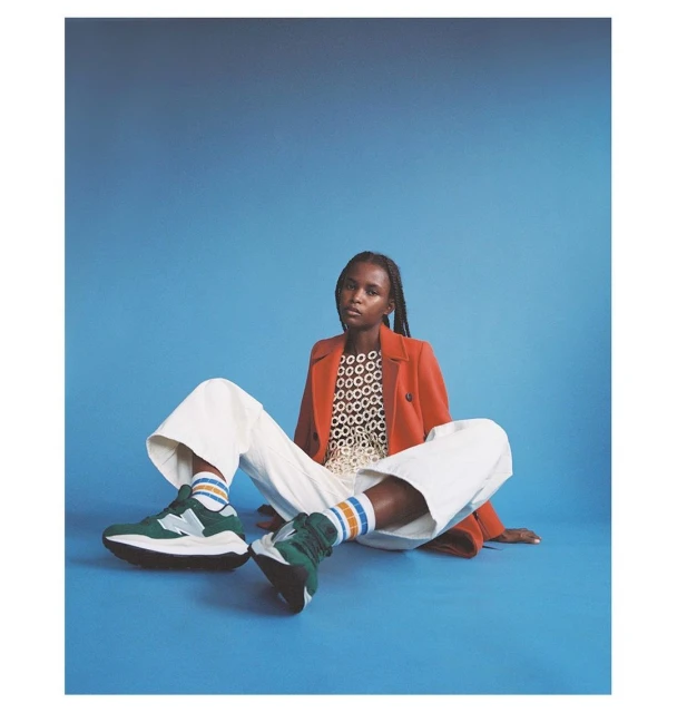 A fashion shot features a woman seated against a blue backdrop, wearing a red blazer, white pants, and green sneakers with colorful socks.