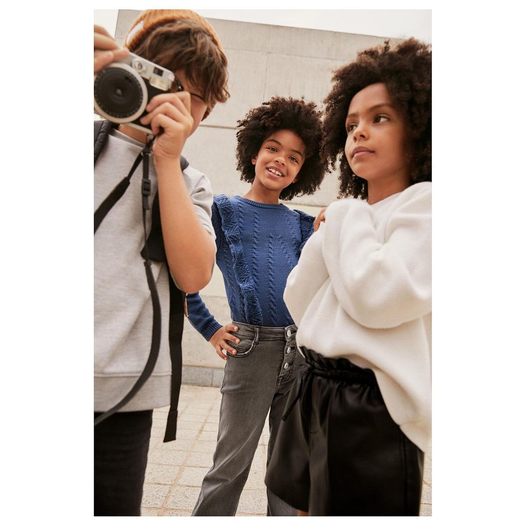 Three young people stand together in a group, one taking a photo with a camera in an outdoor setting during daytime.