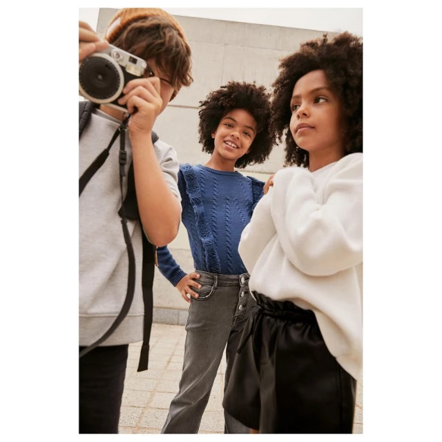 Three young people stand together in a group, one taking a photo with a camera in an outdoor setting during daytime.