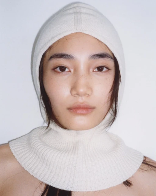 Close-up of a young woman wearing a knitted white balaclava, eyes looking directly at the camera, against a white background.