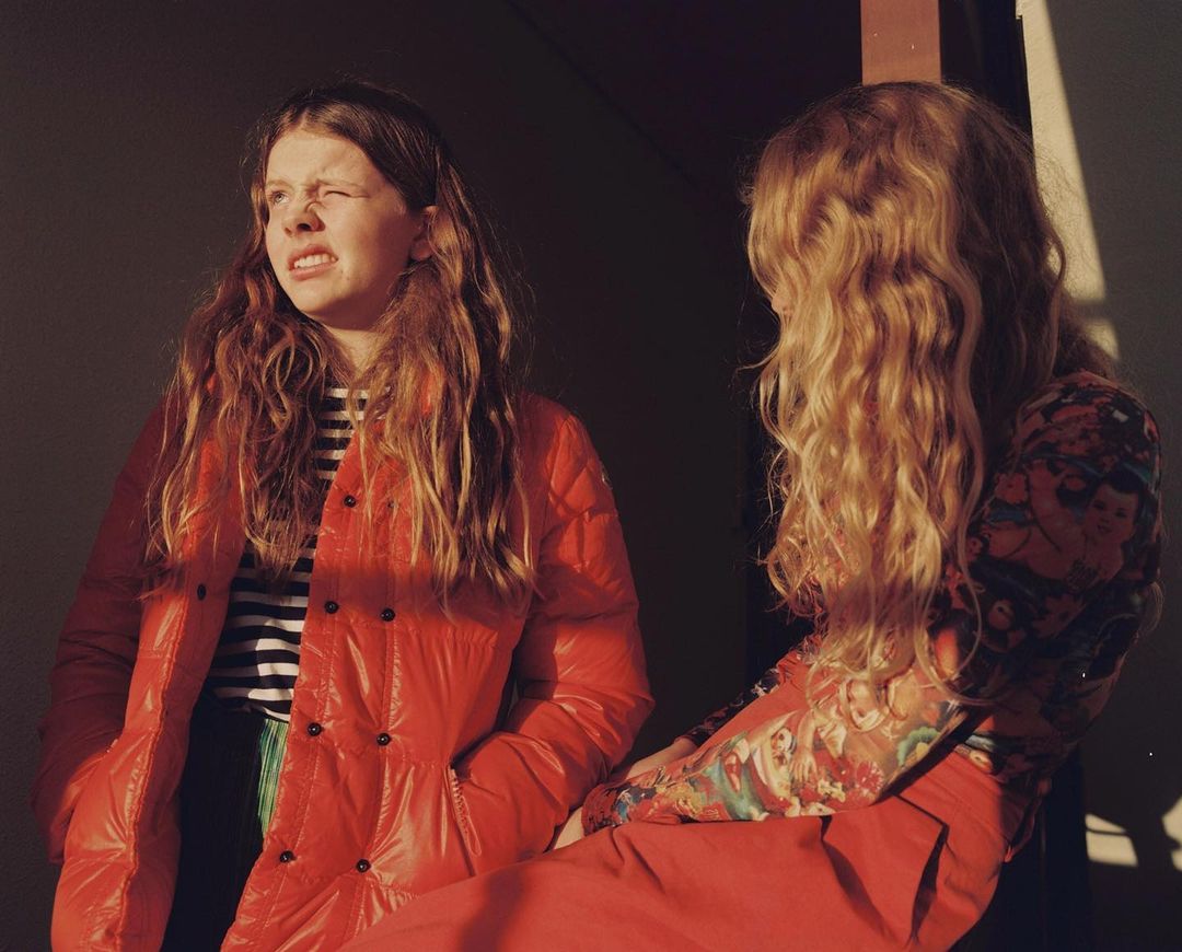 Two women with long wavy hair in an indoor setting. One wears an orange puffer jacket and the other wears a patterned top.