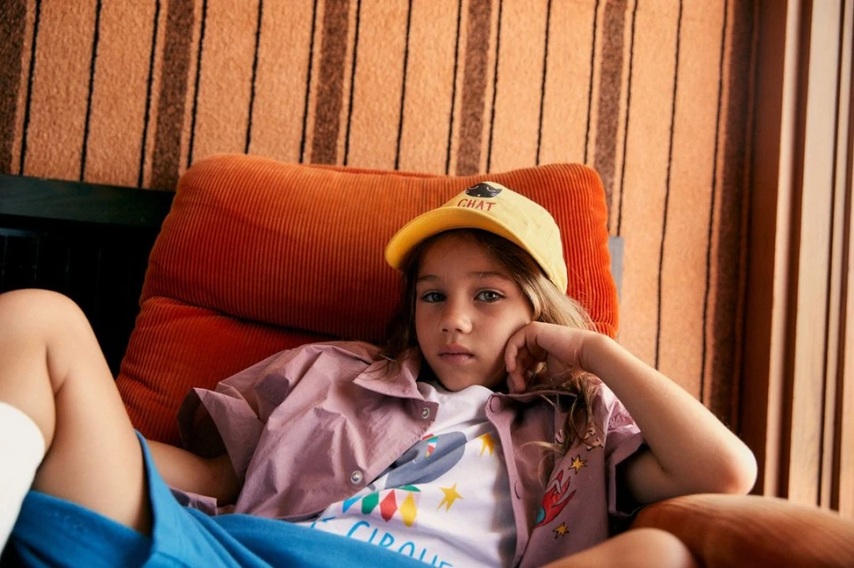 Child model poses in a light purple button-down and blue shorts while relaxing on an orange corduroy chair inside a photo studio.