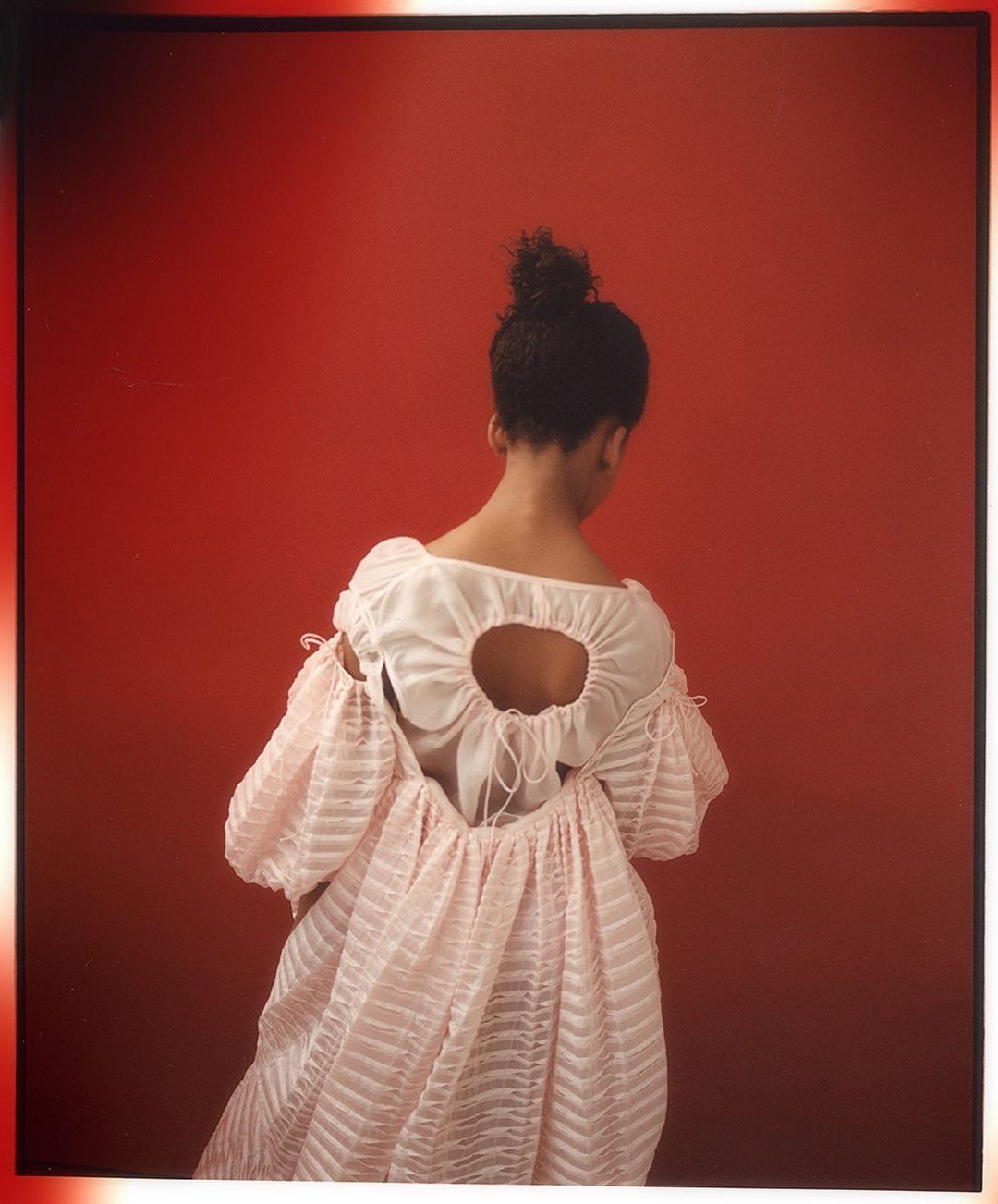 Woman in a pink dress with puffy sleeves poses with her back to the camera against a red backdrop in a fashion photo.