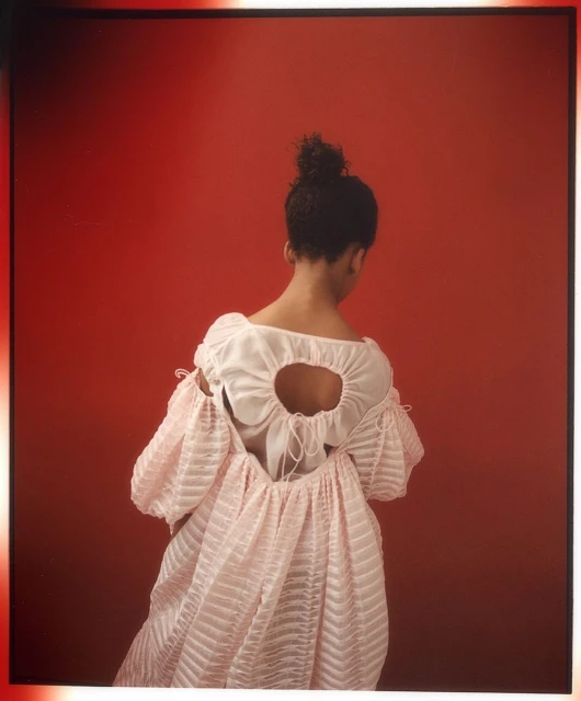 Woman in a pink dress with puffy sleeves poses with her back to the camera against a red backdrop in a fashion photo.