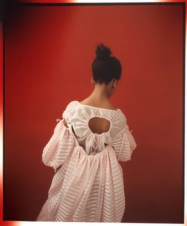Woman in a pink dress with puffy sleeves poses with her back to the camera against a red backdrop in a fashion photo.