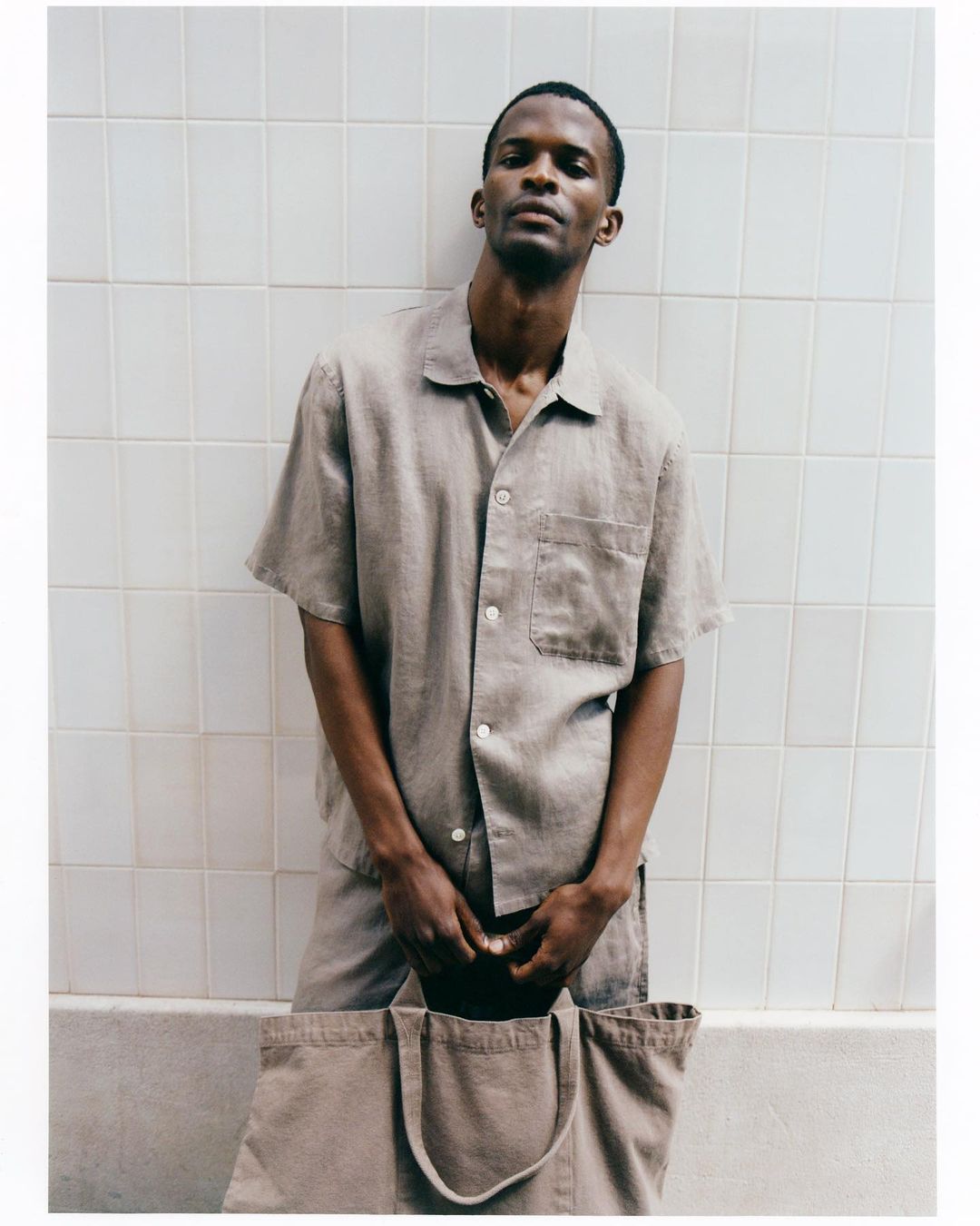 An adult man in a collared shirt and matching shorts holds a tan tote bag against a white tile wall, creating a modern and casual aesthetic.
