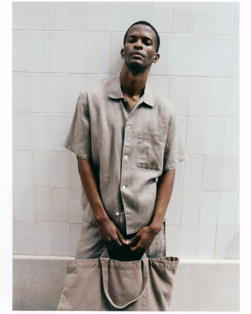 An adult man in a collared shirt and matching shorts holds a tan tote bag against a white tile wall, creating a modern and casual aesthetic.