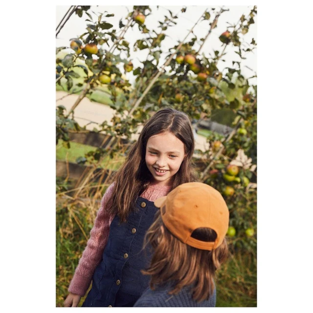 Two young girls near an apple tree with ripe apples, one girl smiling and wearing a blue dress, the other in a cap.