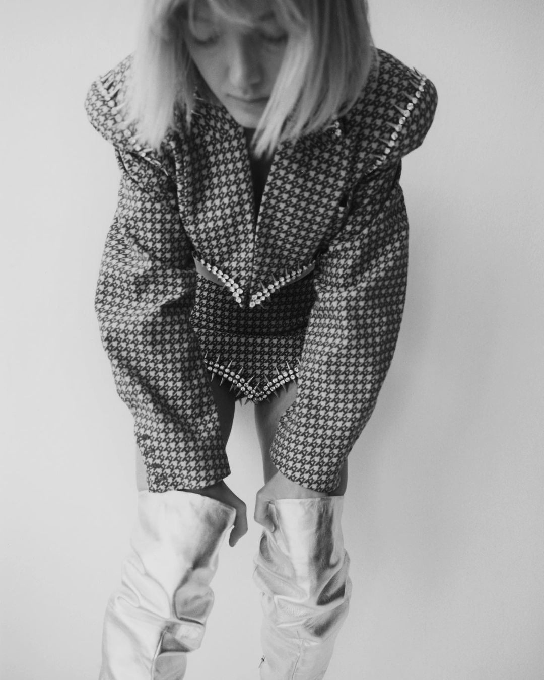 Black and white shot of a woman in patterned jacket, short skirt with stud details, and metallic boots posing against a white backdrop.