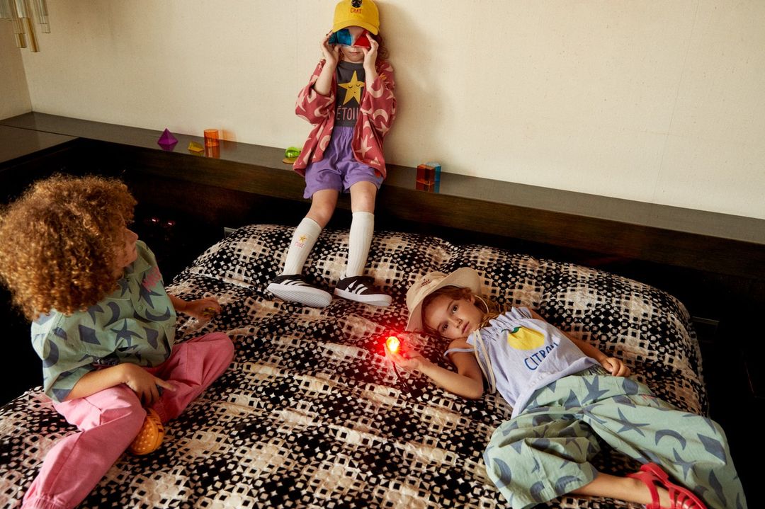 Three children, two girls and one with curly hair, play on a patterned bed with toys, indoor setting, casual style, childhood fun.