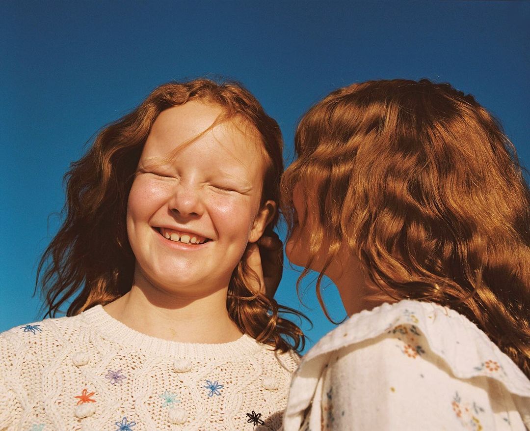 Two young red-haired girls in a close-up portrait against a bright blue sky. One girl is smiling with eyes closed; the other is looking on.