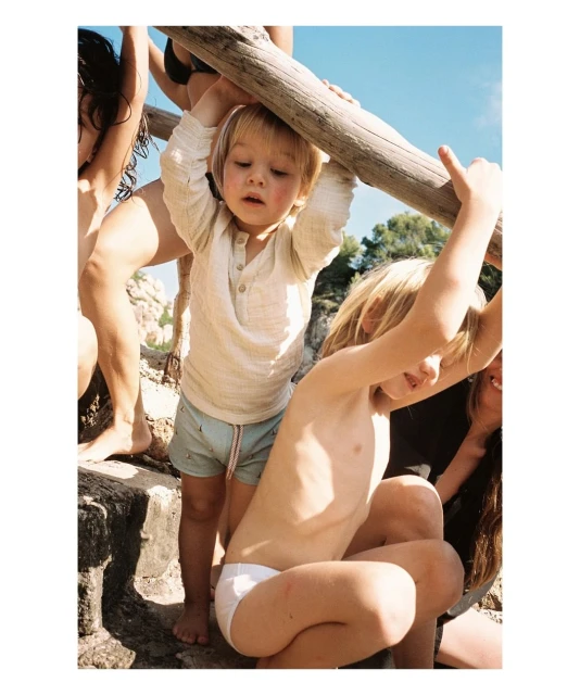 Children are playing on a wooden structure outdoors with a blue sky backdrop. There are multiple children, fair skin, and casual summer clothing.