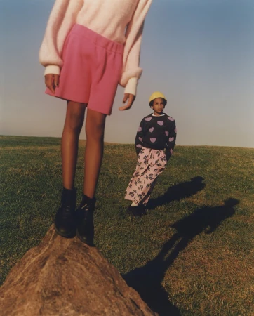 Two children outdoors in a grassy field, one standing on a low rock in the foreground wearing pink shorts.