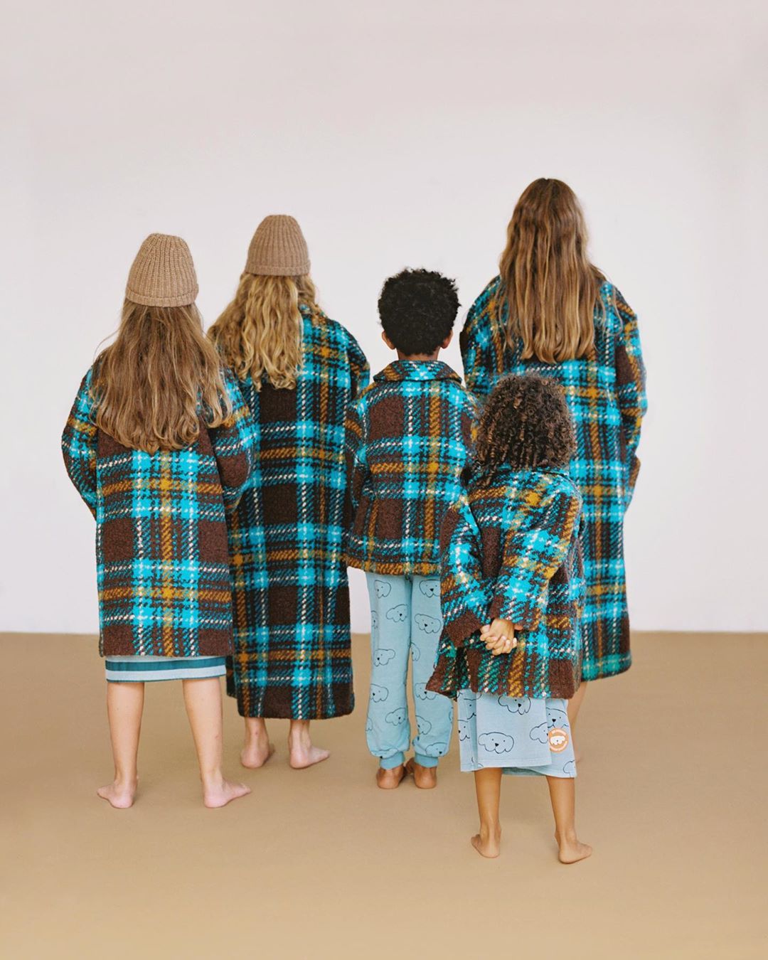 Group of children wearing matching blue-and-brown plaid coats and hats, standing indoors against a neutral background, back view.