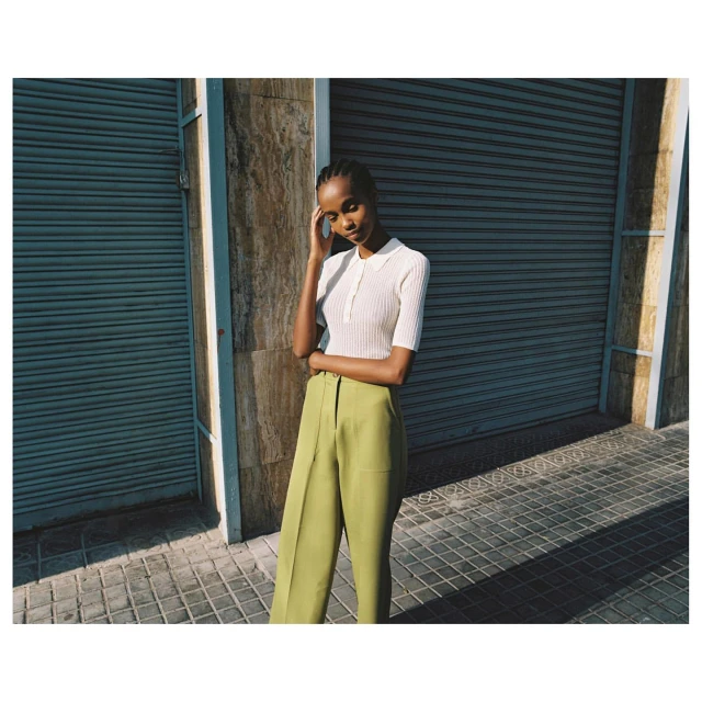 A fashionable dark-skinned woman in a white ribbed shirt and green pants poses against a blue corrugated wall on a paved surface.