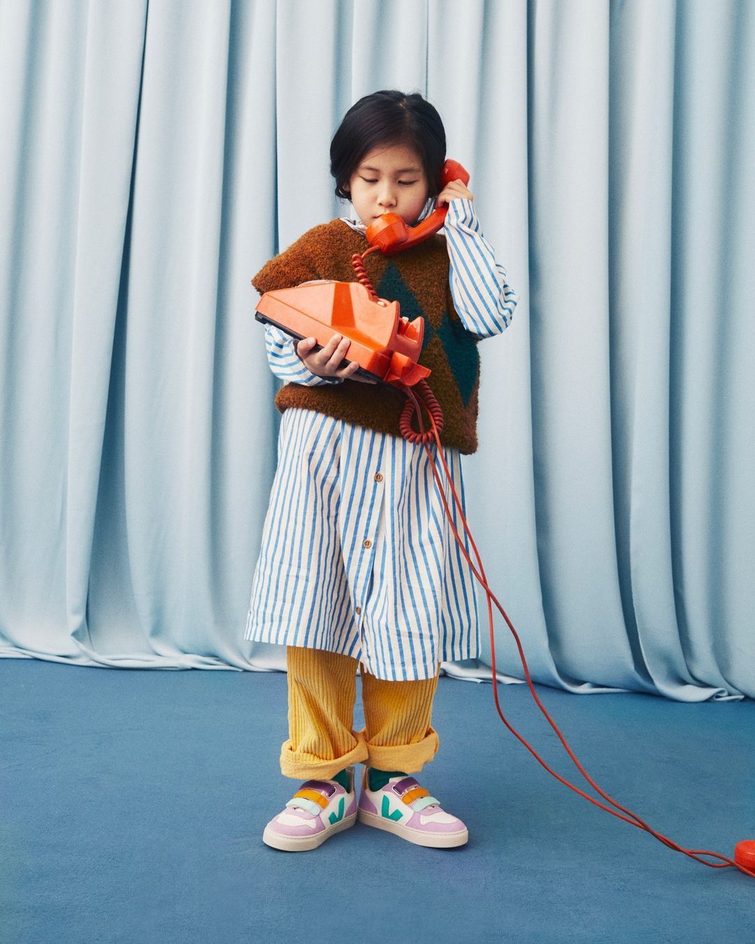 Adorable young girl in striped dress playfully holds an orange retro telephone with a light blue studio backdrop.