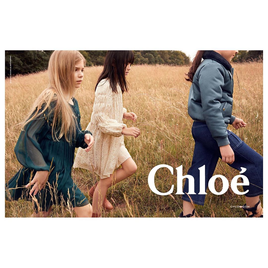 Three young girls walk through a field of tall golden grass wearing casual dresses and a blue jacket, with the brand Chloe in the foreground.