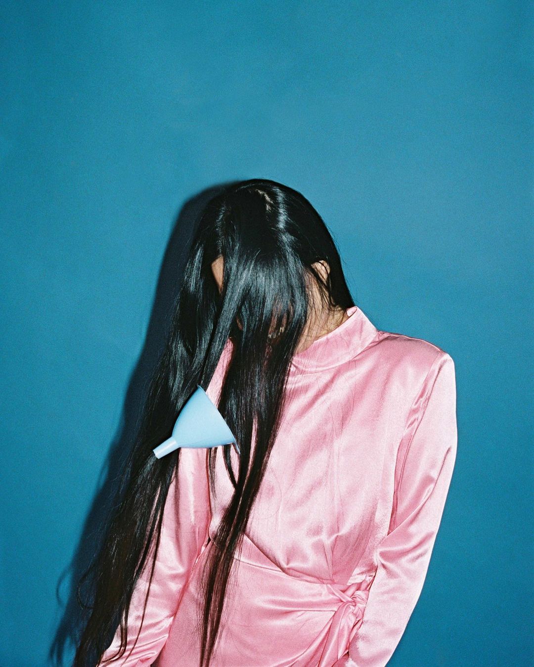 Woman with long black hair poses in a pink dress against a blue background. A blue funnel is playfully placed in her hair.