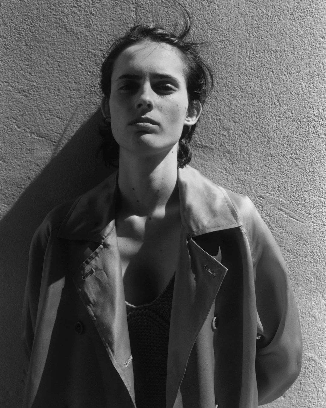 Black and white portrait of a woman wearing a trench coat standing against a textured exterior wall in Paris