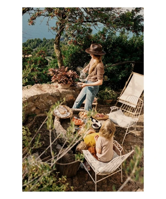 Outdoor picnic scene features a woman pouring water, two children seated nearby, and a table with food, set in a lush garden setting.