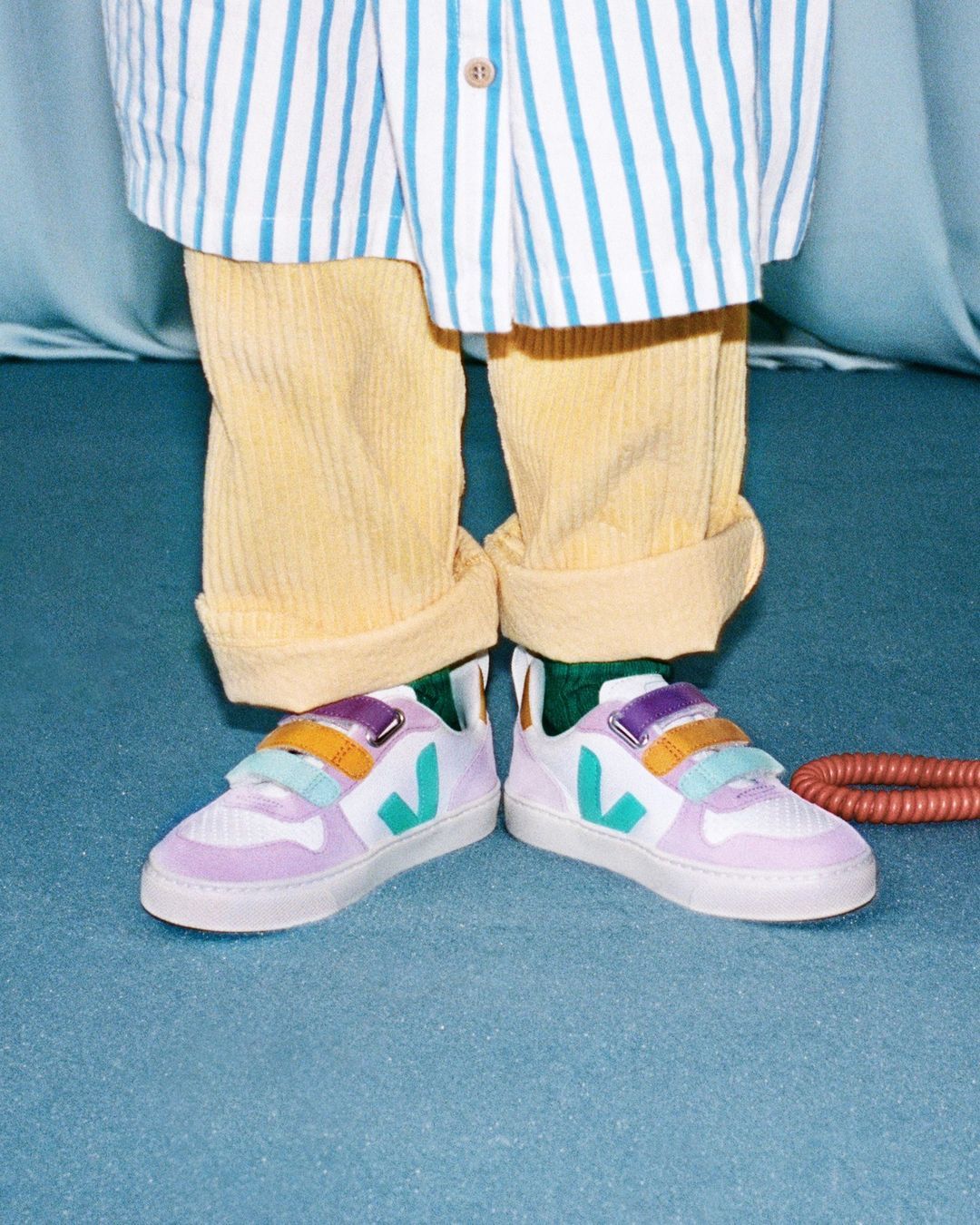 A person is wearing white sneakers with a colored stripe design, yellow corduroy pants, and a striped shirt, showcasing a trendy style.