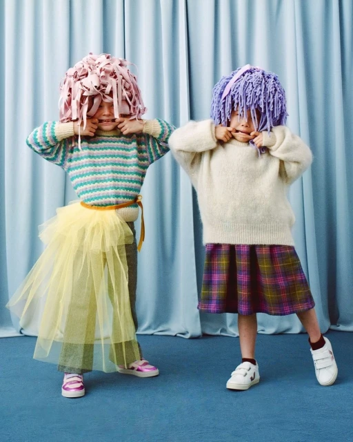 Two children in whimsical paper wigs pose playfully against a blue curtain backdrop, wearing colorful knitwear and skirts.