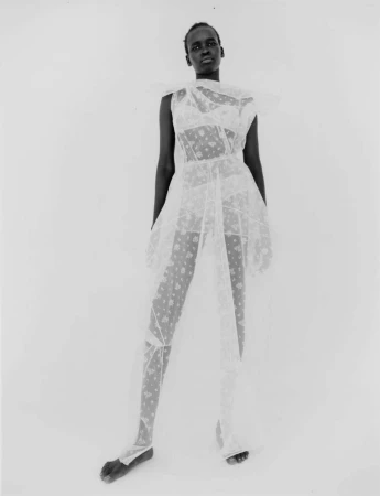 Black and white portrait of a Black female model wearing a patterned sheer dress standing against a seamless background