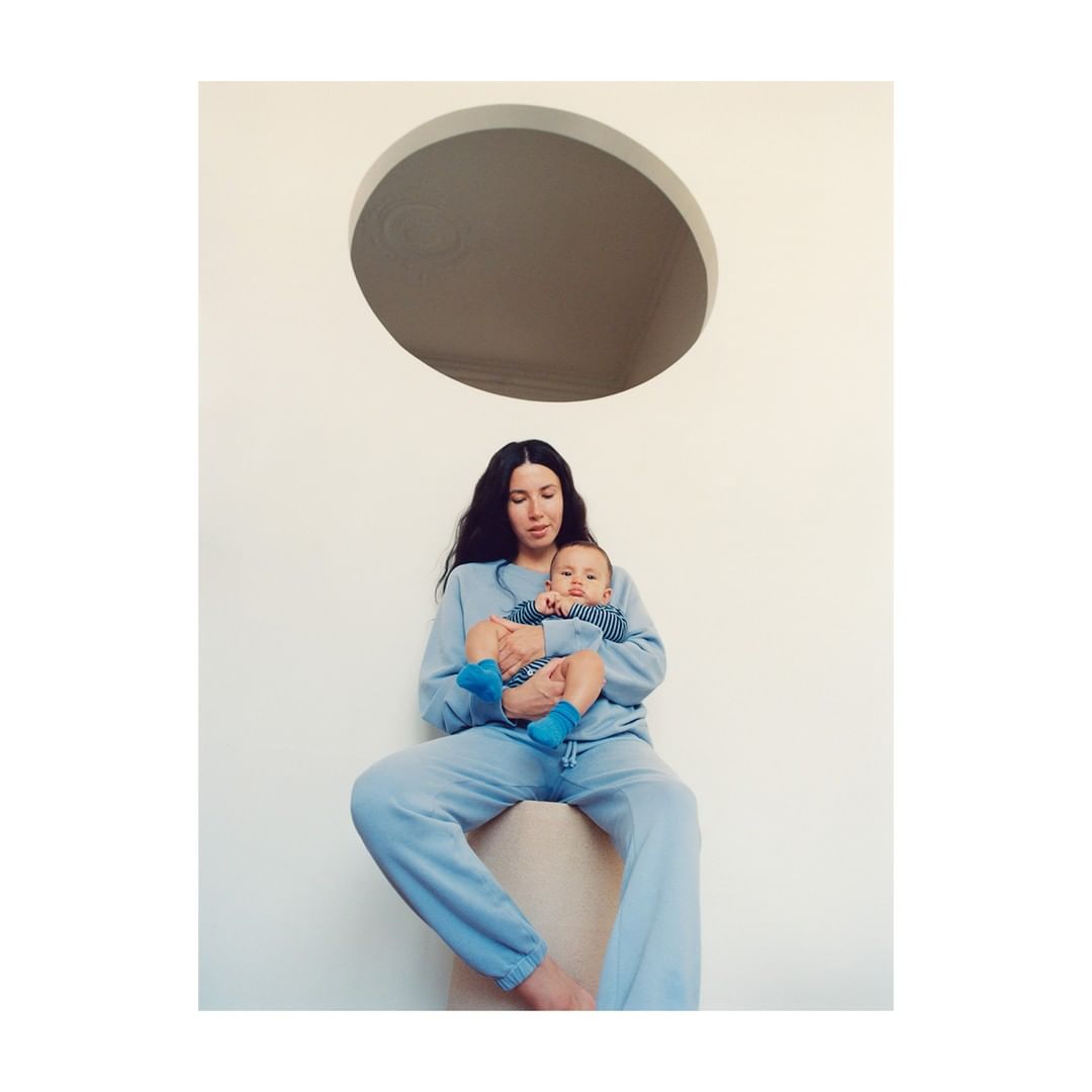 Woman in a blue sweatsuit sits holding a baby in blue socks and striped shirt in front of a white wall with a circular window.
