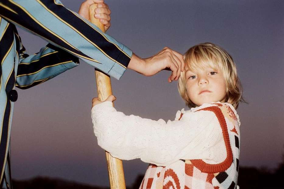Child with blonde hair in a patterned sweater stands next to an adult in a striped jacket, holding a wooden pole against a neutral sky.