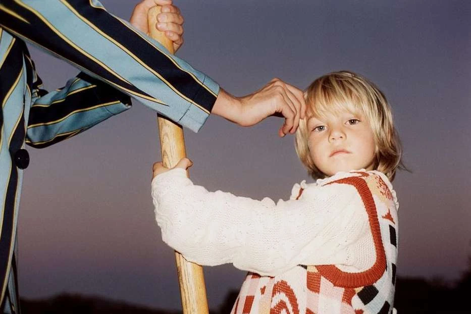 Child with blonde hair in a patterned sweater stands next to an adult in a striped jacket, holding a wooden pole against a neutral sky.