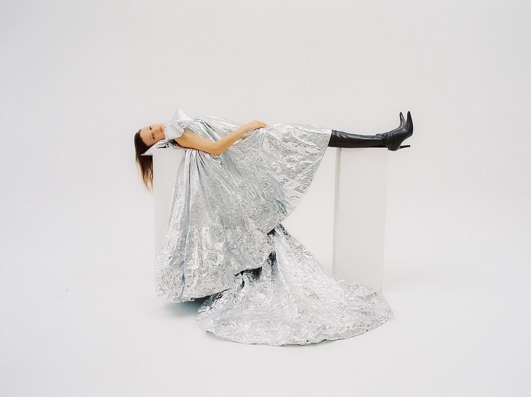 A woman in a reflective silver dress reclines on a white block, wearing black boots against a bright white studio backdrop.