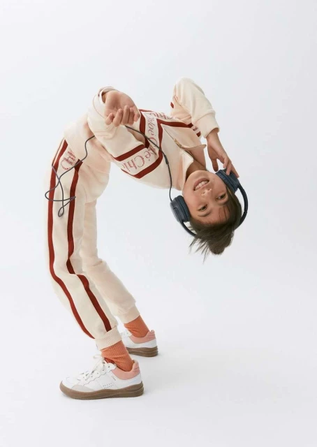 Child bends over backwards in cream-colored sweatsuit with red stripes, wearing headphones against a white background in a studio.