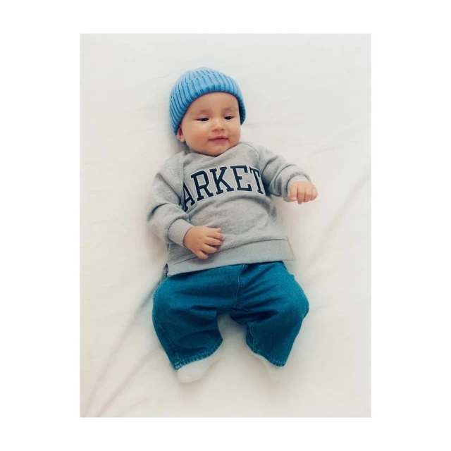 Baby wearing a blue beanie, grey sweatshirt with 'ARKET' printed on it, and blue denim pants lying on a white surface.