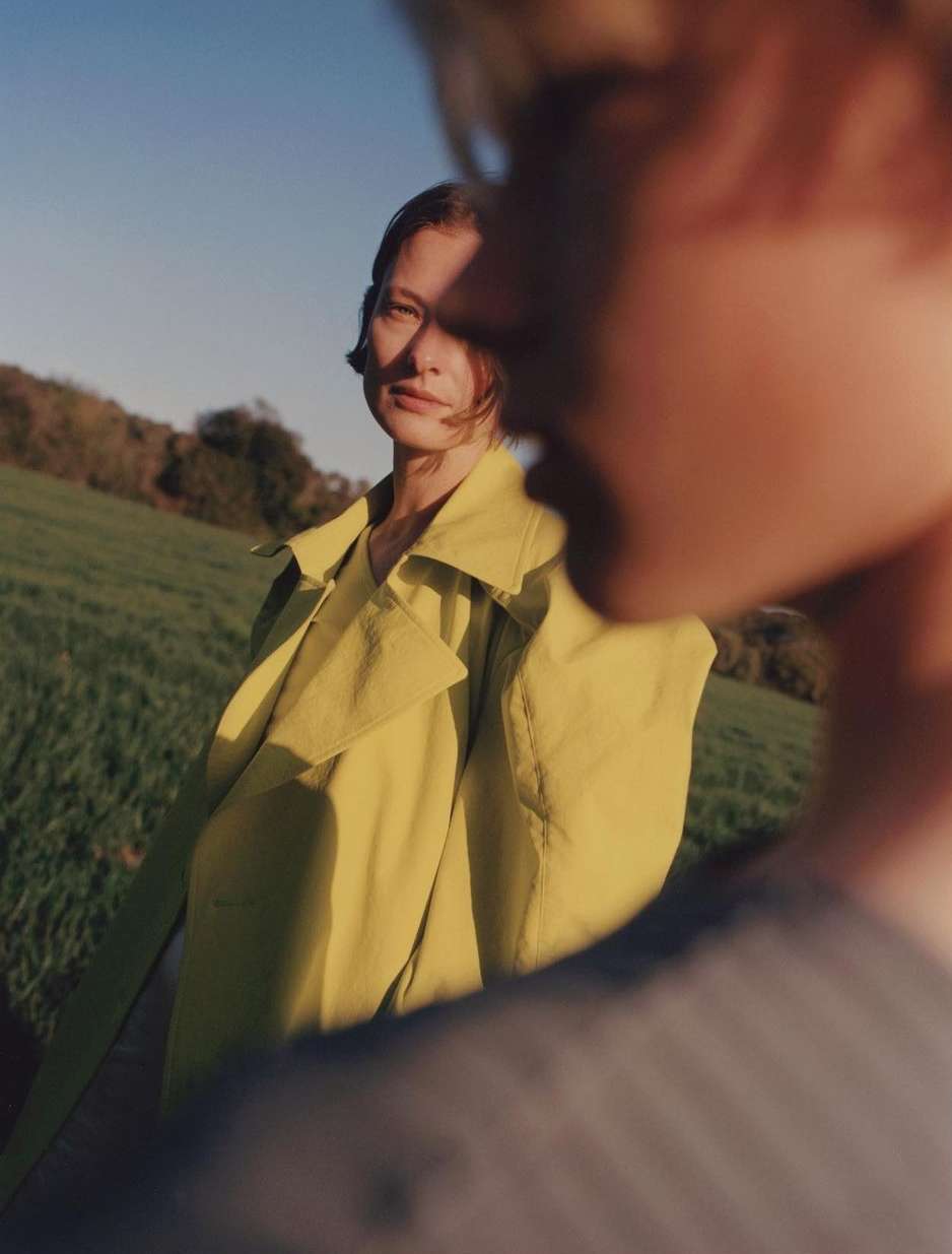 Woman in yellow coat stands in green grassy field under blue sky, viewed behind a blurred figure, creating a unique perspective.